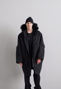 Juodas parka švarkas su kailiniu gaubtu, per didelis dizainas, storas audinys, keli kišenės, užsivilktas ant tamsaus megztinio ir juodų kelnių.