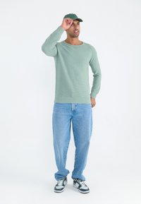 Hellgrüner gestrickter Pullover mit gerippten Bündchen und Säume, kombiniert mit hellblauen, lässig geschnittenen Jeans und schwarz-weißen Sportschuhen.