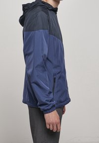 Mörkblå jacka med huva och halvdragkedja, med en kontrasterande mörkblå övre panel, elastiska muddar och sidofickor med dragkedja.