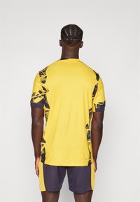 Kollane sportlik jersei tekstuuriga pinnaga, millel on mustad graafilised aktsendid õlgadel ja külgedel. Kombineeritud tumedate lühikestega.