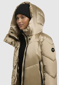 Padded winter jacket in light gold with a large hood, black zipper, and snap buttons. Features a logo patch on the left sleeve.