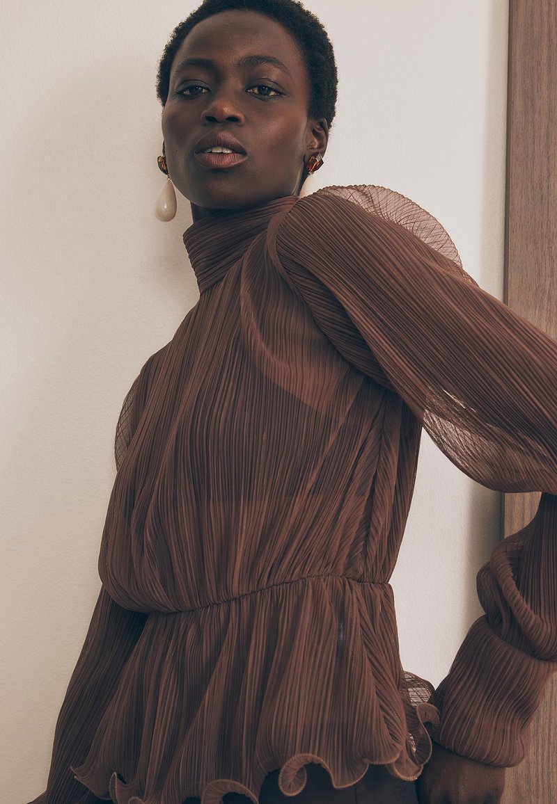 Ruskea laskostettu paita, jossa on läpikuultavat hihat, korkea kaulus ja röyhelöyksityiskohta helmassa. Ominaisuuksiin kuuluu kireä vyötärö ja hillitty tekstuuri.