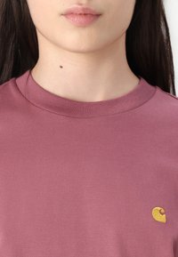 Ljuslila bomulls-t-shirt med en ribbad rundad halsringning och en liten gul broderad logotyp på vänster bröst. Slät textur, avslappnad passform.