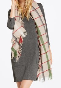 Écharpe à carreaux beige avec des accents rouges et verts, présentant des bords frangés, drapée sur une robe grise. Le tissu semble doux et texturé.