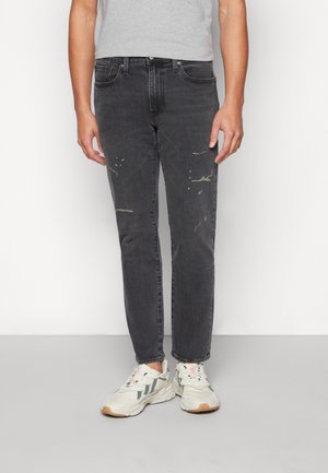 Personne portant un jean noir délavé avec de légères taches de peinture et des baskets blanches avec des touches vertes, debout devant un fond blanc.