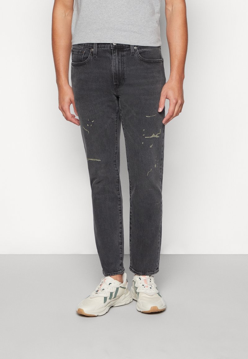 Personne portant un jean noir délavé avec de légères taches de peinture et des baskets blanches avec des touches vertes, debout devant un fond blanc.