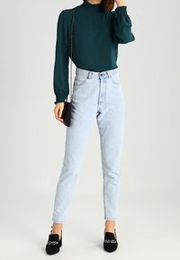 Blusa verde acqua con collo alto e maniche a sbuffo, abbinata a jeans azzurri a vita alta. Scarpe slip-on nere con decorazioni, piccola borsa nera.