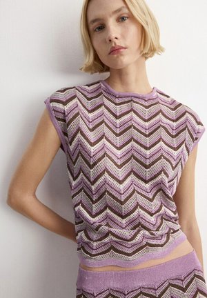 Mujer joven con cabello corto y rubio, vestida con un top de punto sin mangas con patrón de zigzag en púrpura, marrón y blanco, y pantalones a juego.