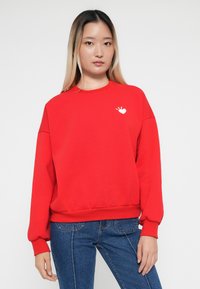 Sweatshirt rouge avec un col rond, des poignets et un ourlet côtelés, présentant un graphisme de cœur blanc sur le côté gauche de la poitrine. Texture douce, coupe décontractée.