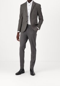 Grijze gestructureerde blazer met bijpassende grijze broek, gedragen over een wit shirt met blauwe knopen, aangevuld met zwarte enkellaarzen.