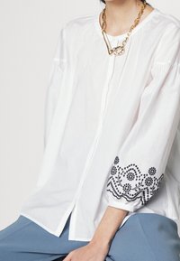 Blouse blanche avec un bouton à l'avant, col rond et motif floral noir brodé sur les manches. Associée à un pantalon bleu et un collier en chaîne dorée.