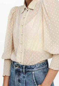 Przezroczysta beżowa bluzka w groszki, z zapięciem na guziki z przodu i bufkami. Posiada elastyczne mankiety oraz klasyczny kołnierzyk. W parze z dżinsami.