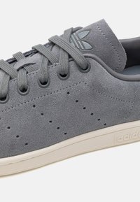 Grå mockasinsneaker med perforerade detaljer, ton-i-ton snörning och vit gummisula. Inkluderar en subtil logotyp på tungan.