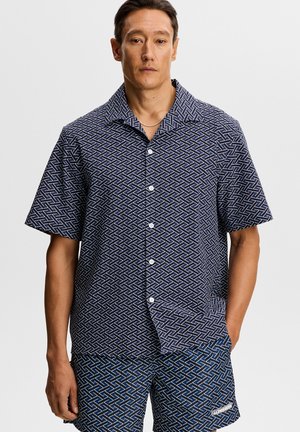 Mann trägt blaues kurzärmeliges Hemd mit Knöpfen und passende gemusterte Shorts mit weißen Zickzacklinien, steht mit einer Hand in der Tasche.