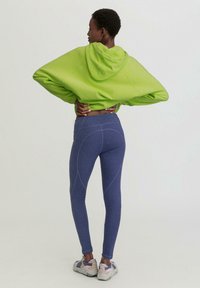 Groene hoodie met een losse pasvorm. Blauwe leggings van een gemêleerd materiaal met contrasterende stiksels. Grijze sneakers met pasteltinten.