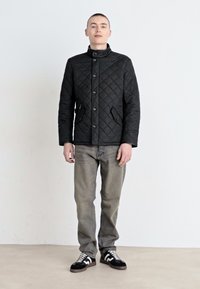 Μαύρο τζάκετ με πλισέ σχέδιο και ψηλό γιακά, με κουμπιά, και πλαϊνές τσέπες; σε συνδυασμό με γκρι τζιν και μαύρα αθλητικά παπούτσια με λευκές λεπτομέρειες.