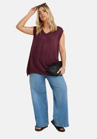 Lila stickad ärmlös topp med V-ringning, kombinerad med vida blå jeans. Modellen håller en svart vävd kuvertväska och har på sig mörka sandaler.