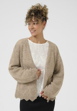 Gilet en tricot beige à coupe ample, manches larges et ourlet festonné. Superposé à un top blanc brodé de fleurs.