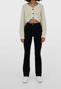 Cardigan court en tricot crème avec trois gros boutons, associé à un jean noir évasé taille haute et des baskets blanches.