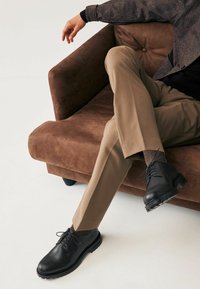 Zapatos de cuero negro con puntas redondas y cordones, llevados con pantalones beige, sentados en un sofá de terciopelo marrón con una textura suave.