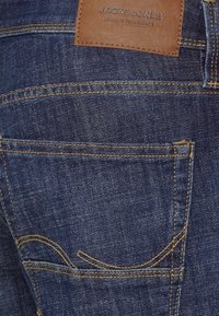 Jeans in denim blu scuro con cuciture gialle a contrasto, dotati di una toppa in pelle marrone in vita e dettagli sottili sulle tasche posteriori.
