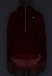 Red hooded jacket with a metallic zipper and logo; paired with orange patterned shorts, featuring zippers on the sides.