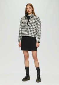 Zwart-wit houndstooth jas met een gekraagde halslijn, knoopsluitingen en lange mouwen, gecombineerd met een zwarte rok en zwarte laarzen.