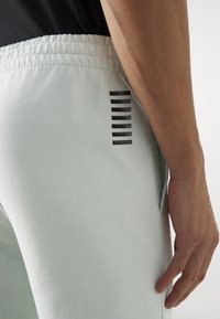 Detailní záběr na dolní část trupu a ruku osoby v světlešedých teplácích s černým vodorovným pruhem poblíž kapsy.