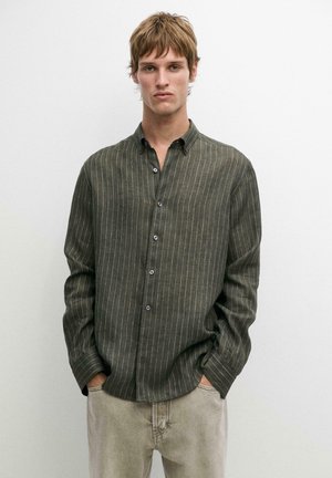 Junger Mann mit kurzen hellbraunen Haaren, der ein dunkelgrünes, Nadelstreifen-Knopfhemd und hellbeige Hosen trägt, die Hände in den Taschen.