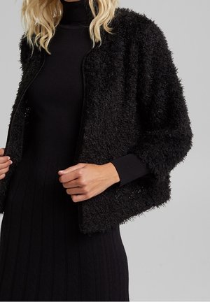 Femme portant une veste noire duveteuse ouverte sur une robe noire à col roulé côtelée, debout devant un fond gris uni.