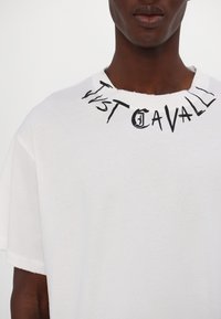 Bílá bavlněná tričko s kulatým výstřihem, s černě potisknutým logem "Just Cavalli" kolem límce. Měkká textura a volný střih.