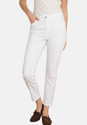 Femme portant un jean blanc taille haute et ajusté, une main dans la poche, et des mocassins marron à pampilles, sur fond blanc.