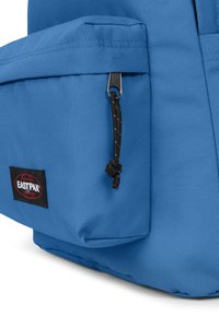 Sininen kangaskassi, jossa on etutasku, musta vetoketju ja Eastpak-logopatch. Rakenteeltaan teksturoitu materiaali ja kestävä muotoilu.