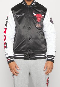 Chaqueta varsity en blanco y negro con un logo de toro rojo, múltiples parches, botones a presión y un cuello y puños de canalé a rayas.