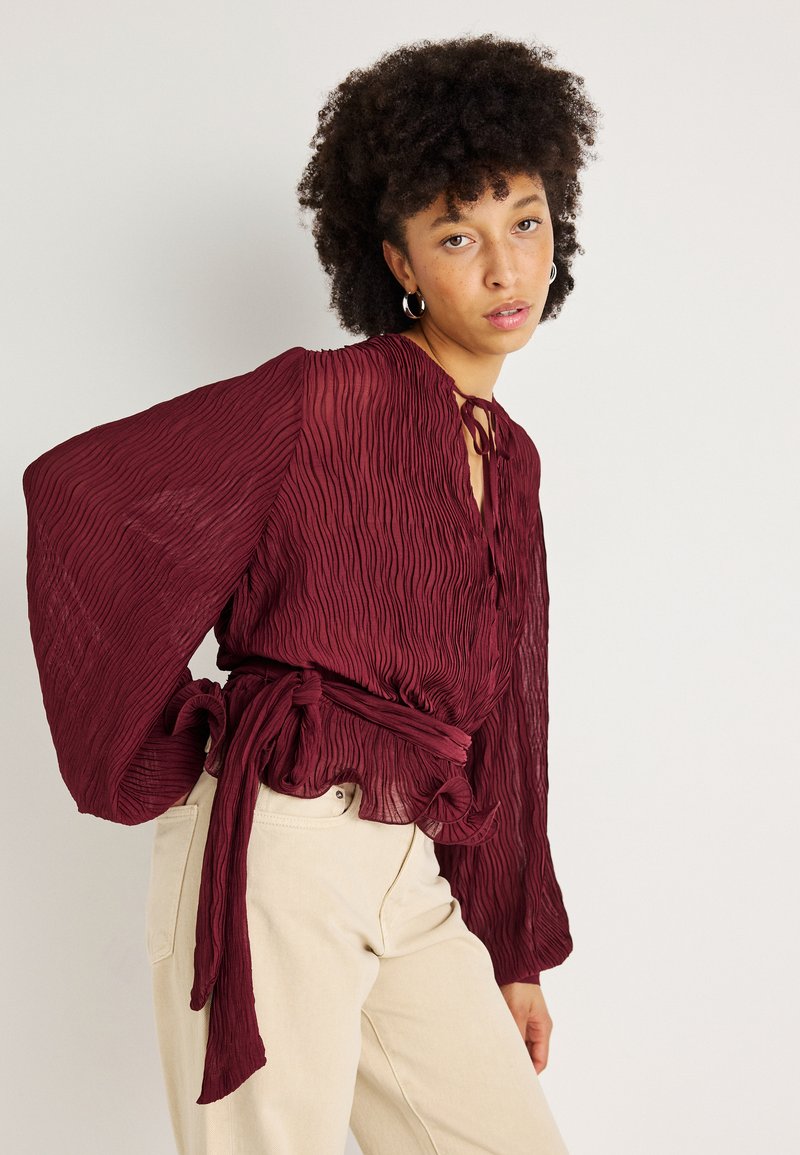 Blusa burgundy plissada com mangas balão, laço na frente e cintura ajustada. Combinada com calças bege. Tecido texturizado com um design fluido.