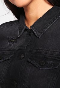 Chaqueta de mezclilla negra con cuello clásico, detalles desgastados y cierre de botones. Incluye bolsillos en el pecho con solapa y botones decorativos.