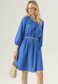 Vestito blu a pois con maniche lunghe a sbuffo e vita stretta, dotato di una cintura beige. La modella tiene una piccola borsa a tracolla beige.