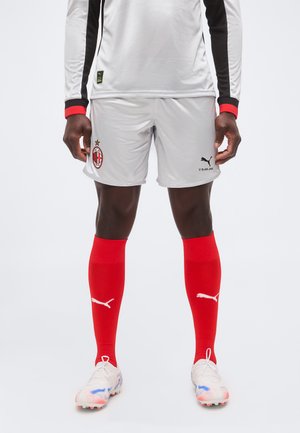 Άτομο που φοράει γκρι ποδοσφαιρικά σορτς AC Milan, κόκκινες κάλτσες Puma, λευκά παπούτσια με τάπες και μακρυμάνικη γκρι φανέλα με μαύρες και κόκκινες λεπτομέρειες.