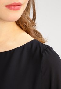 Haut en tissu noir à la texture lisse, présentant un large décolleté et un drapé doux aux épaules, mettant l'accent sur une élégante simplicité dans le design.