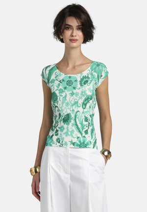 Žena v zelenej a bielej kvetinovej krátkosleevej blúzke v kombinácii s bielymi vysokými nohavicami a zlatými náramkami.