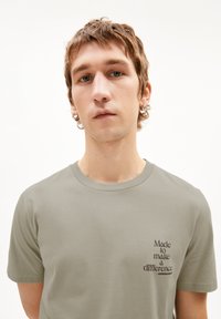 Olijfgroen katoenen T-shirt met korte mouwen en een ronde hals; heeft een bedrukte slogan op de linker borstpartij in donkerbruin.