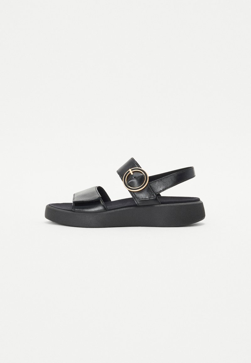 Black leather sandals with a flat platform sole, strap across the instep, and a circular metal accent on the side. Textured detailing on straps.