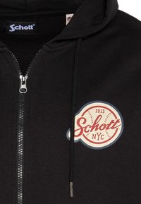 Sudadera con capucha negra y cremallera, con cordones. Presenta un logo circular en rojo y crema que dice "Schott NYC" con motivos de béisbol. Textura suave.