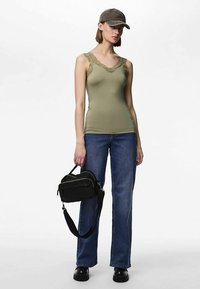 Olívazöld ujjatlan póló csipkés szegéllyel, kék farmerrel párosítva. A modell fekete, több cipzáras rekesszel rendelkező táskát tart, és szürke sapkát visel.