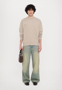 Beige sweatshirt with a crew neckline, paired with faded blue wide-leg jeans, holding a brown leather shoulder bag. Plain background.
