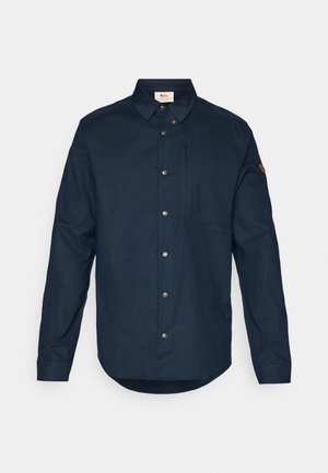 Chemise bleu marine à boutons, à manches longues, avec un col pointu et une poche poitrine unique. Elle se distingue par des boutons métalliques et un logo sur la manche.