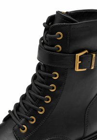 Bottine noire en cuir avec lacets, œillets dorés et sangle à boucle sur le côté.