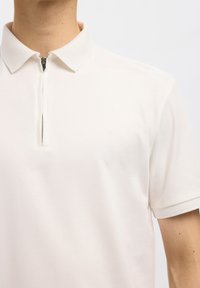 Mann trägt ein schlichtes weißes Kurzarm-Poloshirt mit halb durchgehendem Reißverschluss vorne und strukturiertem Kragen sowie Ärmelsäumen.
