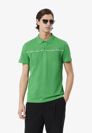 Polo verde in cotone, con colletto, maniche corte e una striscia bianca in accentuazione orizzontale sul petto, con un logo sul lato sinistro.