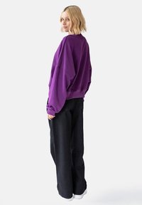 Fioletowy, oversize'owy sweter z opadającymi ramionami, grubymi mankietami i ściągaczem na dole, w połączeniu z szerokimi czarnymi spodniami i białymi trampkami.